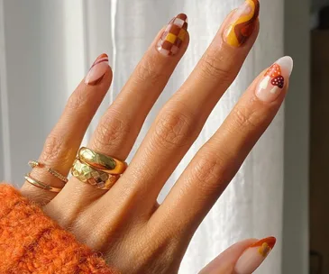 Hand with autumn-themed nail art featuring swirls, mushrooms, and plaid on long nails; wearing gold rings.