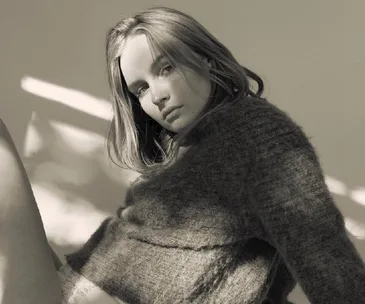 Sepia-tone portrait of a woman in a cozy sweater, seated thoughtfully against a softly lit background.