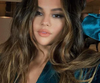 A woman with long wavy hair and soft makeup poses, wearing a teal satin outfit.