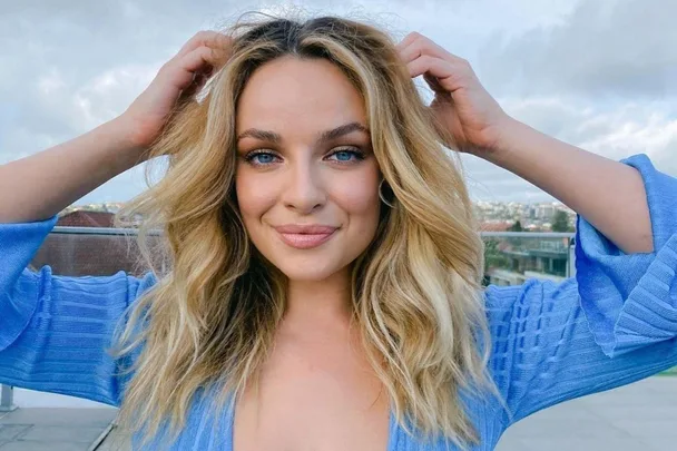 Young woman with long blonde hair, smiling in a blue top, standing outdoors with a cityscape background.