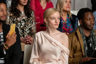 A woman in an elegant off-the-shoulder dress sits among a diverse crowd at a fashion event, looking focused.