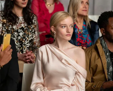 A woman in an elegant off-the-shoulder dress sits among a diverse crowd at a fashion event, looking focused.