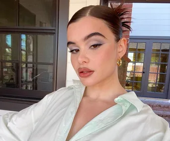 Woman with sleek hairstyle and hoop earrings, wearing light makeup, poses in front of glass doors and a window.