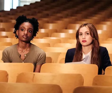 Two women sitting in a lecture hall with wooden seats, one in a green top and the other in a suit, both appearing focused.