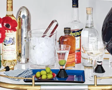 A stocked drinks trolley with bottles, cocktail shaker, ice bucket, glasses, olives, red flowers, and napkins.