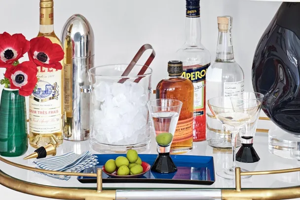 A stocked drinks trolley with bottles, cocktail shaker, ice bucket, glasses, olives, red flowers, and napkins.