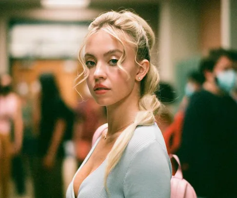 Cassie from Euphoria with blonde bubble braids, looking over her shoulder in a school hallway.