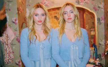 Two women with long blonde hair in matching blue cardigans stand side by side in front of a floral wallpaper.