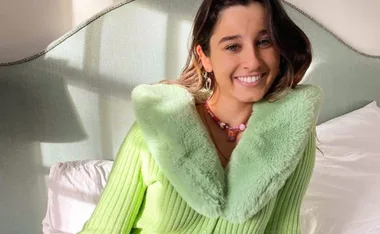 Woman smiling, wearing a light green furry-collared sweater, sitting on a bed in a sunlit room.