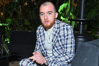 Bearded man in a plaid suit sitting, hands folded, surrounded by lush greenery and ambient lighting.