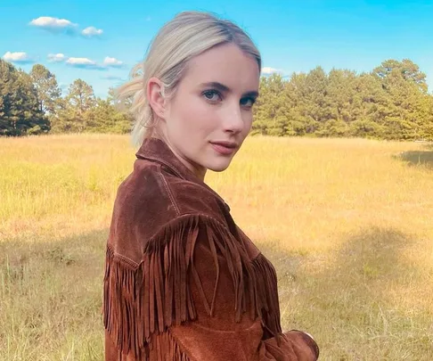 Actress in a brown fringe jacket stands in a sunny field with trees and blue sky in the background.