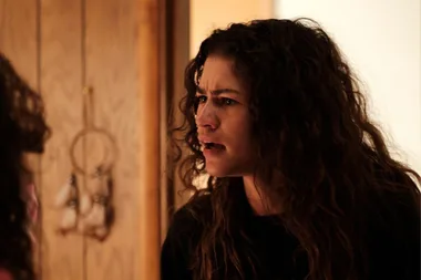 A character in "Euphoria" with curly hair looks intense and emotional in a warmly-lit room.