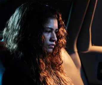 Rue sits somberly in a car, deep in thought, with her curly hair illuminated by dim light.