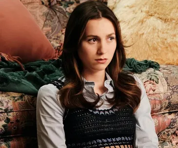 A woman with brown hair and a serious expression sits on a floral-patterned couch with pillows and a blanket.