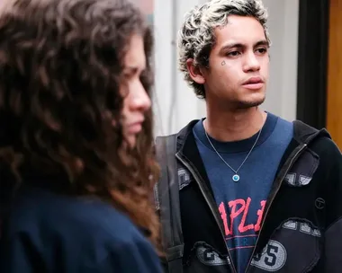 A young man with blond hair and a teardrop tattoo looks ahead, while a woman with curly hair stands blurred in the foreground.