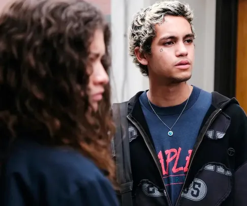 A young man with blond hair and a teardrop tattoo looks ahead, while a woman with curly hair stands blurred in the foreground.