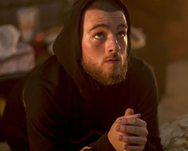 A bearded man in a hoodie kneels with clasped hands, gazing upward in a dimly lit room.