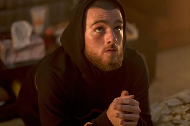 A bearded man in a hoodie kneels with clasped hands, gazing upward in a dimly lit room.