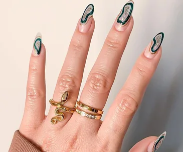 Hand with geode nail art in black and silver, wearing a gold snake ring and two gold bands.