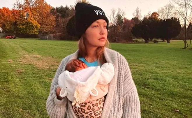 Woman in a beanie holding a baby outdoors with autumn trees in the background.