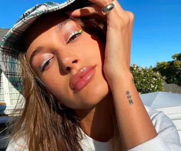 Close-up of a woman with glossy skin and rainbow eyeliner, wearing a plaid hat and sunlight illuminating her face.