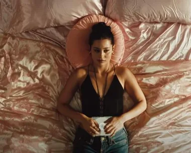 A woman lies on a bed with pink satin sheets, wearing a black top and jeans, holding an iPod.