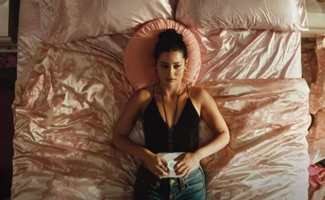 A woman lies on a bed with pink satin sheets, wearing a black top and jeans, holding an iPod.