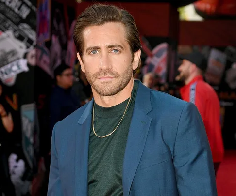 A man in a blue suit and green shirt stands on a red carpet at an event with people in the background.