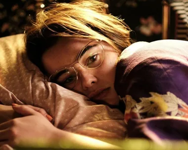 A person with short hair and glasses lies on a pillow, covered with a patterned blanket, looking contemplative.