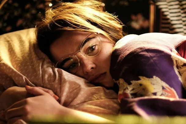 A person with short hair and glasses lies on a pillow, covered with a patterned blanket, looking contemplative.