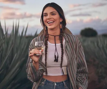 Smiling woman in a field, holding a glass of tequila, wearing a striped shirt and braids.