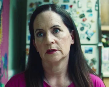 A woman with long brown hair and pink top stands in a kitchen with a fridge covered in magnets behind her.