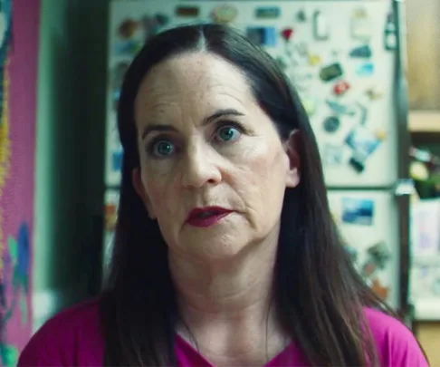 A woman with long brown hair and pink top stands in a kitchen with a fridge covered in magnets behind her.