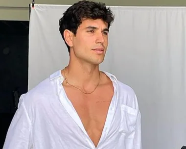 A man in a white open shirt poses confidently against a plain backdrop.