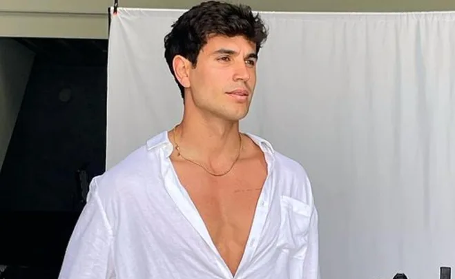 A man in a white open shirt poses confidently against a plain backdrop.