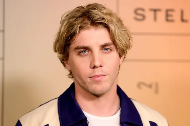 Man with blond hair and blue eyes in a jacket stands against a beige backdrop with text in the background.