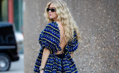 Woman with long wavy hair wearing sunglasses and a blue patterned dress with an open back, standing outdoors.