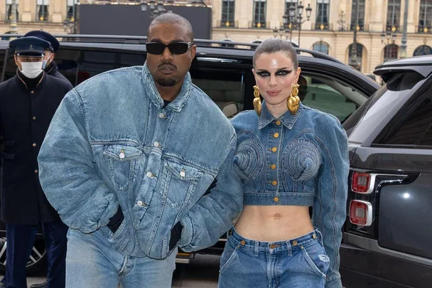 Two individuals in matching denim outfits stand outside a vehicle, wearing sunglasses and stylish earrings.