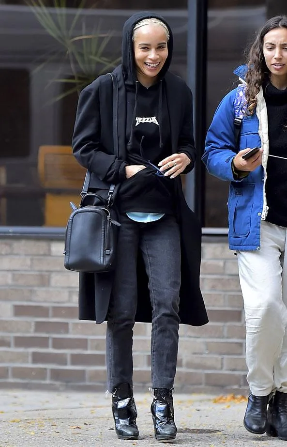 A woman in a black hoodie and jeans walking outside, carrying a black bag, smiling with a friend.