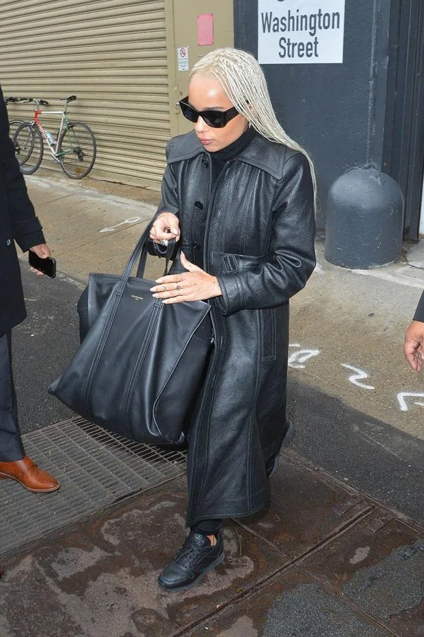 Person in black coat and sunglasses walking on street with large black bag.