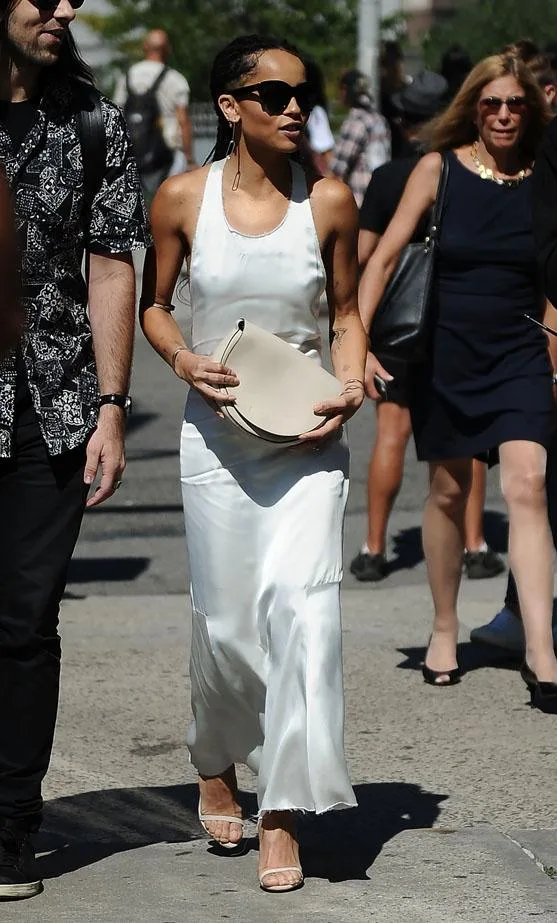 Zoe Kravitz in a white sleeveless dress and sunglasses, holding a beige clutch, walking with others on a sunny day.
