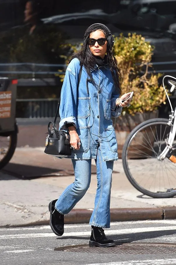 Woman in double denim, sunglasses, and headphones crossing street, holding a phone with a black purse.