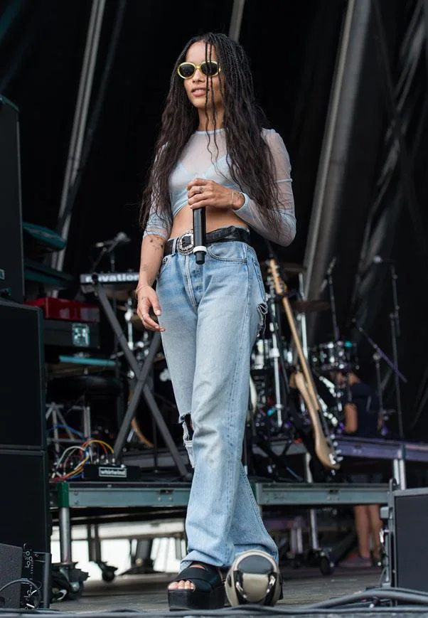 Singer in sunglasses and casual attire holding a microphone on stage, with musical instruments in the background.