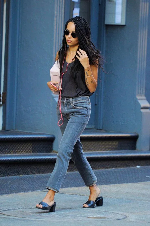 Young woman with long braids, sunglasses, and casual outfit walks down a city street listening to headphones.
