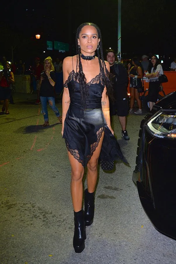 A person in a black lace dress and boots walking on a street at night, surrounded by people and a car.