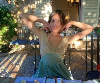 A woman in a lace top poses with her arms up, seated outdoors at a table, sunlight filtering through leaves.