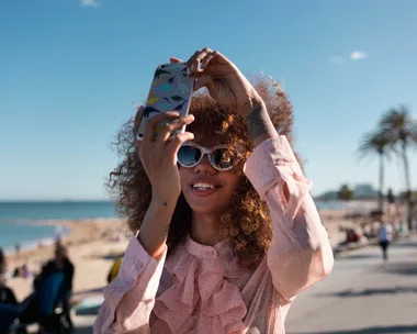 A woman with curly hair and sunglasses takes a selfie on a sunny beach promenade.