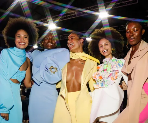 Models in vibrant, colorful outfits joyfully pose together under bright lights at a fashion event.