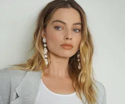 A woman with long wavy blonde hair wearing a light gray blazer, white top, and pearl earrings.