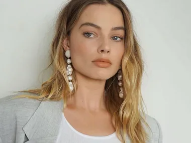 A woman with long wavy blonde hair wearing a light gray blazer, white top, and pearl earrings.